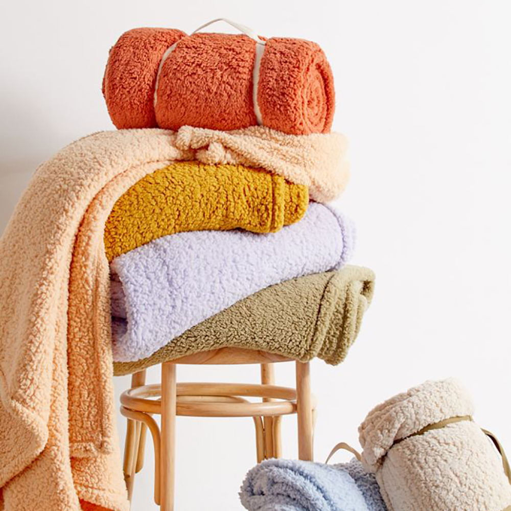 Photo of a stack of jewel-tone and pastel-colored fuzzy blankets on top of a stool and on the ground.