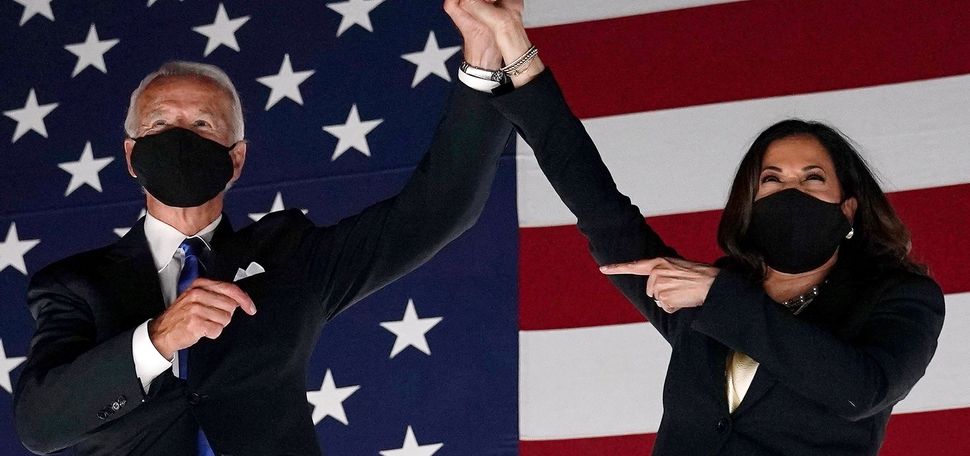 A photo of president-elect Joe Biden and Vice President-elect Kamala Harris clasping hands and raising them above their heads. They are standing in front of an American flag. Both are wearing masks.