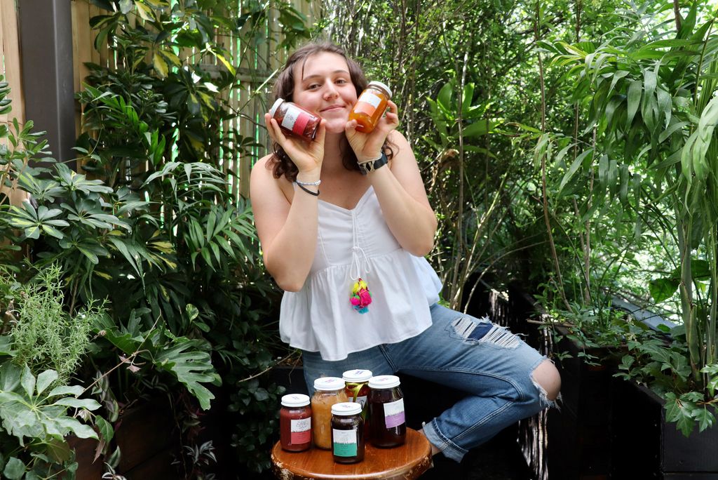 A photo of Inés Santacruz holding jars of jams and preserves up in front of her face. Several jars also sit on a table in front of her.