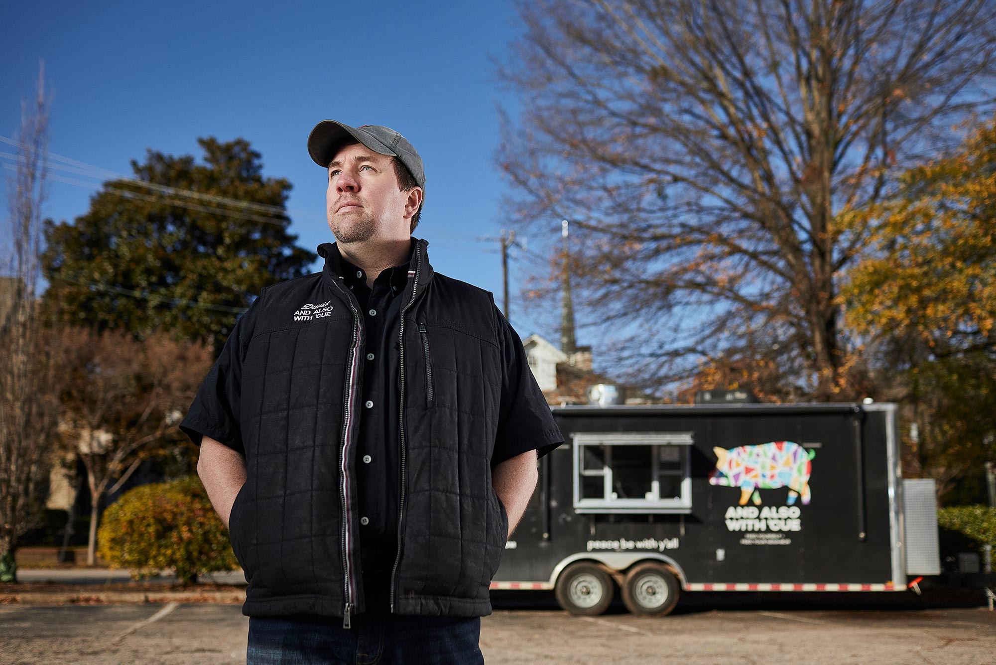 Photo of David With in a black vest, shirt, and baseball cap, standing with his hands in his pockets and looking off to the side. His the black trailer where he runs his BBQ business out of is behind him; blue sky and trees are seen in the background.