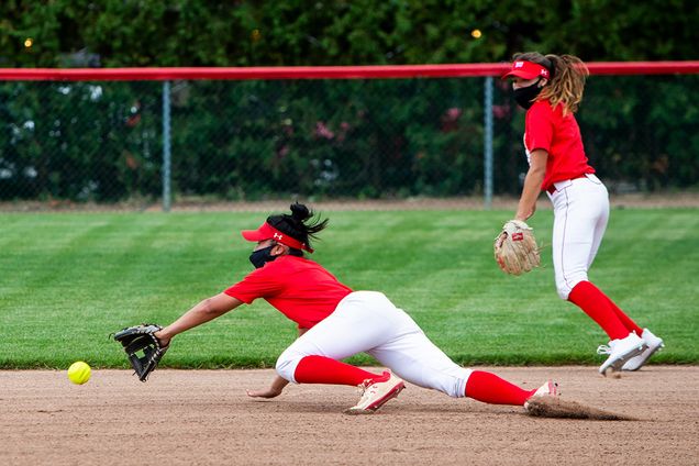 Photo of two of BU softball players, both wearing black masks and red and white uniforms. One woman lunges to the ground to field the ball while another backs her up.
