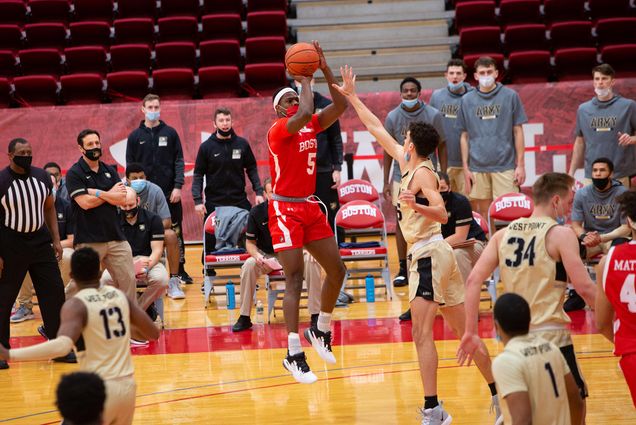 Photo of BU men’s basketball player Walter Whyte (CAS’22) mid-air as he shoots a basketball in front of a player on the opposing team, who tries to the block the shot with his hand. The crowded basketball floor foregrounds the shot, while coaches and people on the bench are seen in the background.