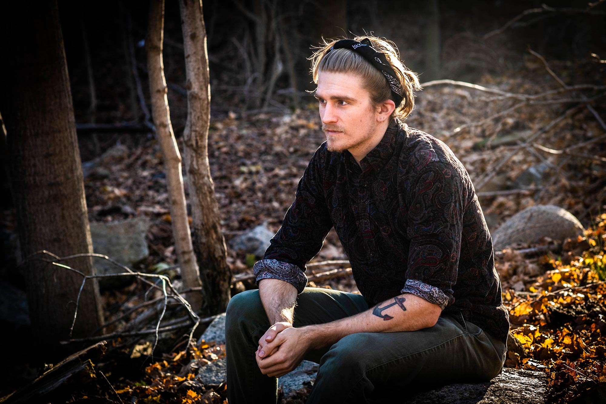 Photo of Macken Murphy (CAS’20) sitting on a rock outside and staring of into the middle distance. He wears a black headband, black button down, and has a tattoo of an anchor on his left arm.