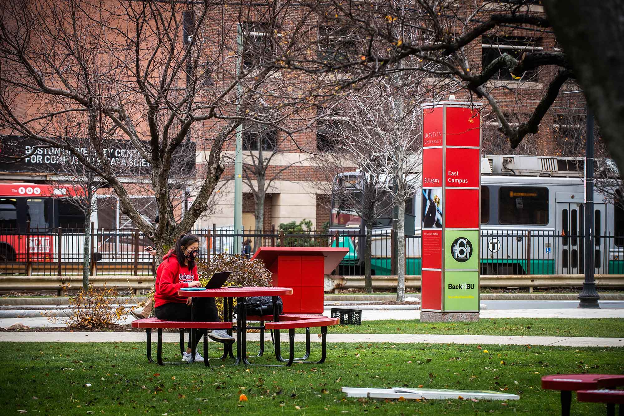 A young female student in a red sweatshirt and black face mask sits by herself at a red picnic table on a lawn on BU's Campus. A signpost behind her reads "Back2BU"; the green line and a BU shuttle bus drive by in the background. The trees are bare.