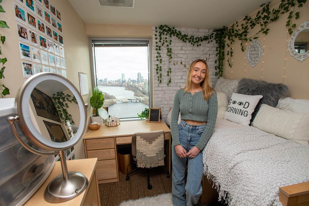 Photo of Casey Evans (COM’21) standing in her StuViII quad with a million-dollar view of Boston’s skyline in the window at the back of the room. The dorm room is beautifully decorated with hanging plants, a wall of photos, and a bed with many pillows, one of which reads “good morning.”