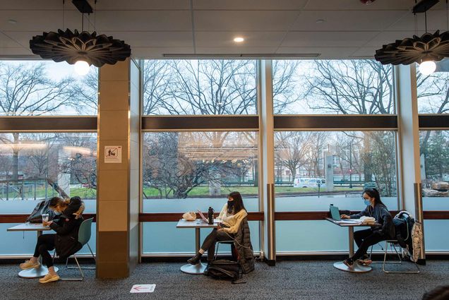 Photo of three students sitting at single person tables as they study in front of their laptops with face masks on. The bare trees of the BU Beach are seen through the windows behind them.