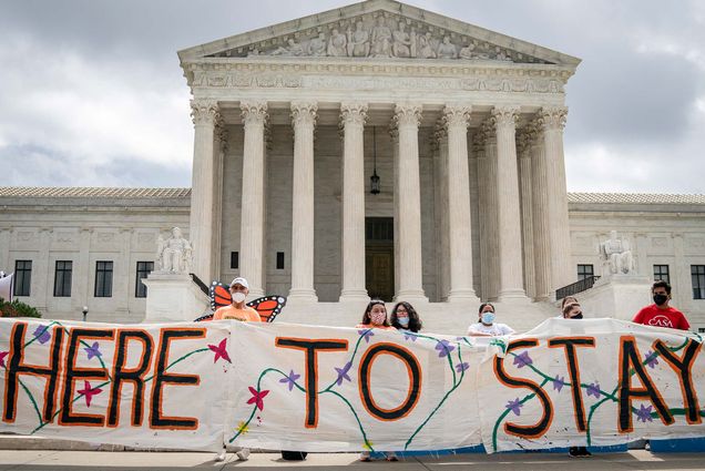 Photo taken outside of the US Supreme Court on June. In the image, DACA recipients and their supporters hold a painted cloth sign that reads “HERE TO STAY” that is decorated with a floral pattern. One of the people present wears a pair of monarch butterfly wings.