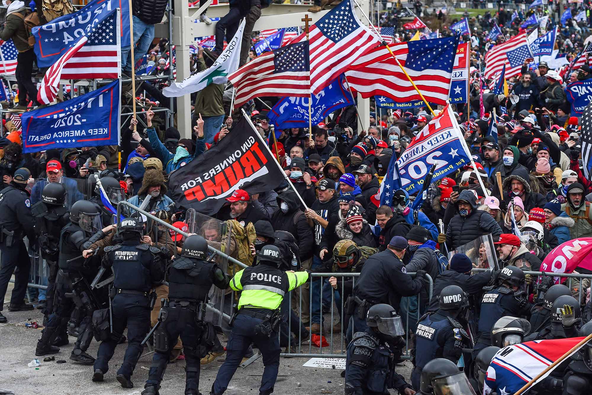 Photo of Trump supporters clashing with police and security forces as they storm the US Capitol in Washington, DC on January 6, 2021. Police officers, dressed in riot gear, push back a metal barrier as insurgent Trump supporters holding American flags and Trump 2020 flags try to break through the line.