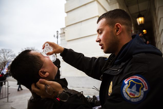 Photo of a pro-Trump protester being tended to by a police officer while suffering the effects of chemical agents used to disperse crowds after protesters stormed the grounds the Capitol Building on January 6, 2021 in Washington, DC. The police offer holds the mans head gingerly and pours water down his face; neither man is wearing a face mask.