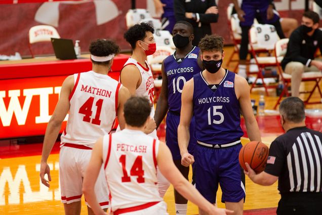 Photo of BU and Holy Cross men's basketball players walking by one another during the pre-game of one of their opening season games. Each of young men wear a face mask, and a referee with a ball in his hands is seen in the foreground.