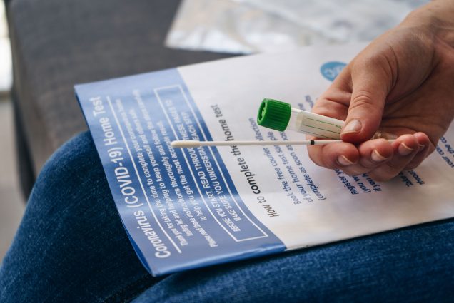 Photo of a person sitting with an at-home COVID test and booklet in their lap. They hold a swab and a tube; the booklet is labelled “coronavirus (COVID-19) Home Test”.