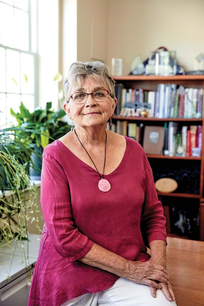 Potrait of Bonnie-Jean Brooks in a pink blouse and white pants sitting in her office; a book case is seen in the background.