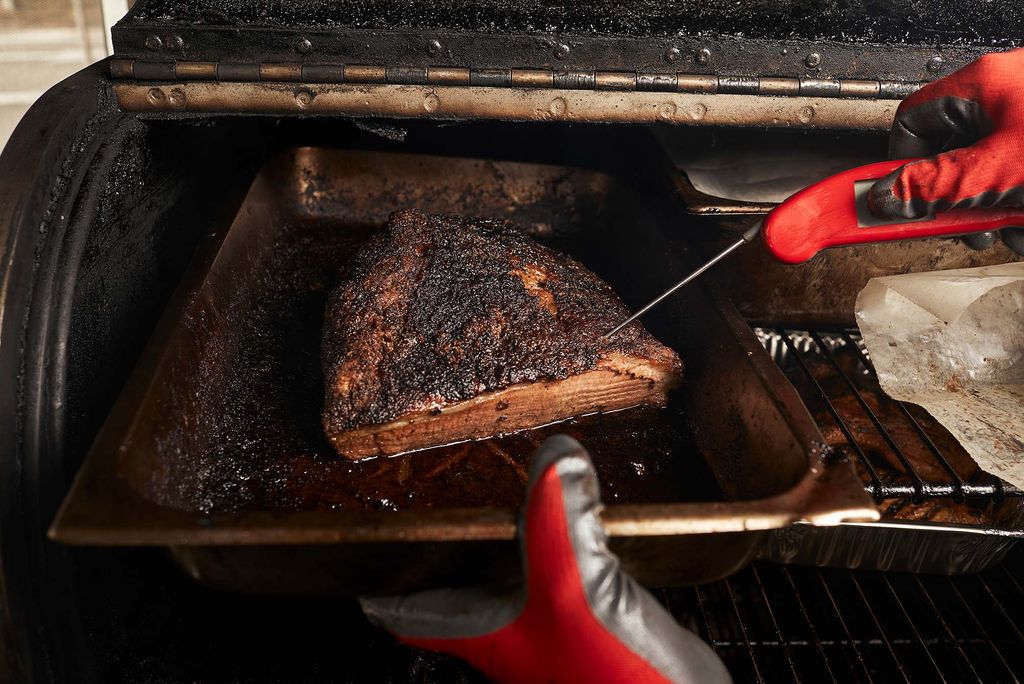 Photo of a pair of gloved hands using an electronic thermometer to take the temperature of a piece of Texas Style Brisket. The brisket is crispy, juicy and brown; the oven around it is greasy and blackened. 