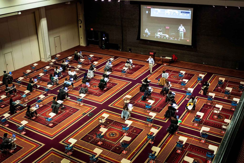 Photo taken from the catwalk of the Metcalf Ballroom as Professor Igor Lukes teaches in a blue button down and khakis at the front of rows of students spaced 6 feet apart. A camera is set up and Lukes is seen on a projector behind him. Everyone wears face masks.