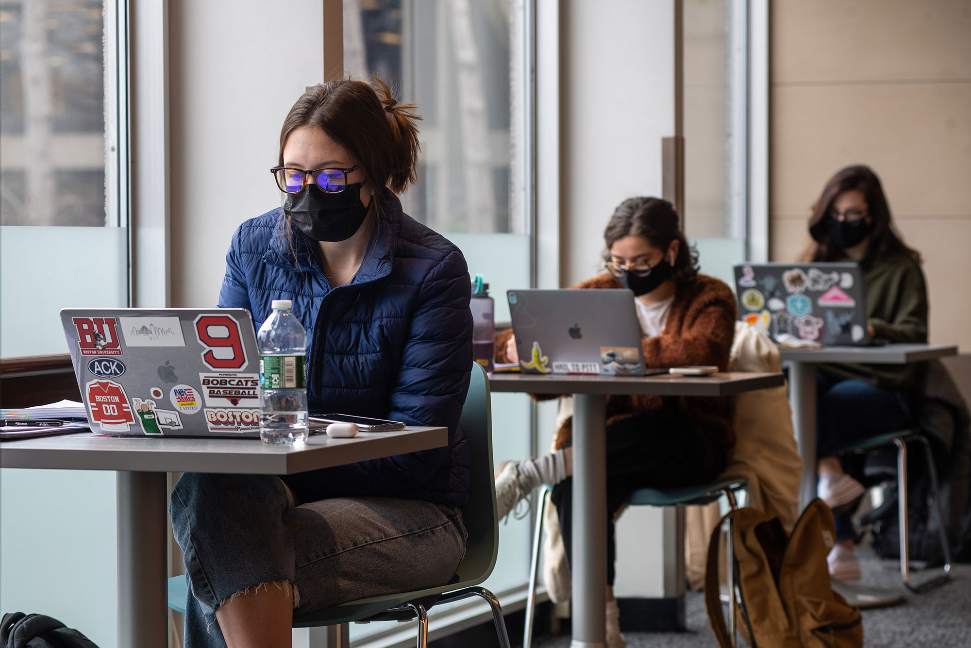 Photo of three students with masks, sitting at separate, one-person tables in the GSU in a row. Each student looks at her laptop and attends lecture virtually.