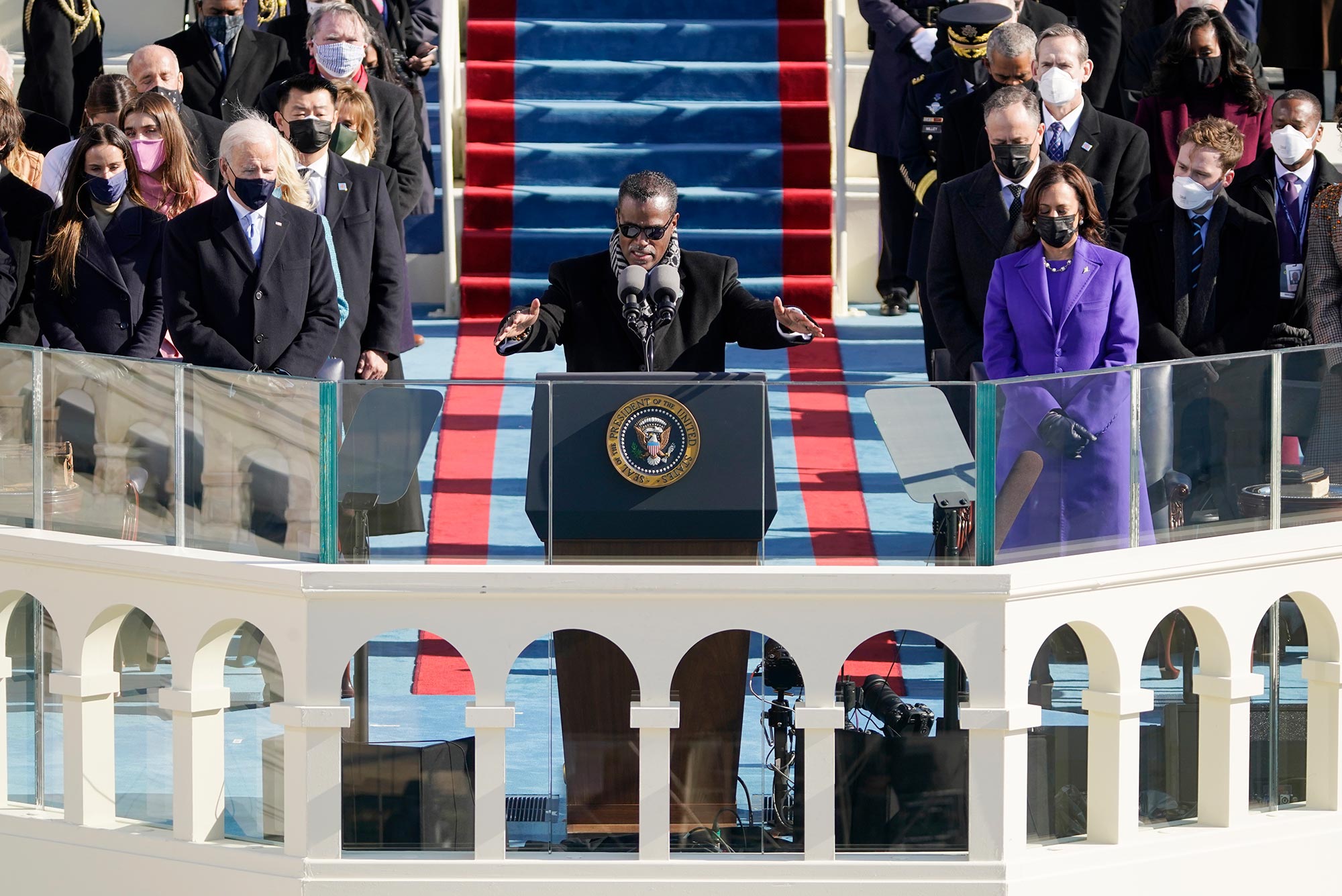 Photo of Rev. Silvester Beaman giving the benediction during the 59th Presidential Inauguration at the U.S. Capitol in Washington, Wednesday, Jan. 20, 2021. He wears a black jacket and sunglasses and extends his arms forward as he speaks from the podium; he is flanked by Biden and Harris.