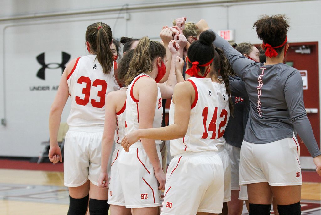 Photo taken from behind the women’s basketball team as they huddle raise their hands all raised towards the center of the huddle. Some women wear white jerseys while others wear gray long sleeves; they all wear face masks.