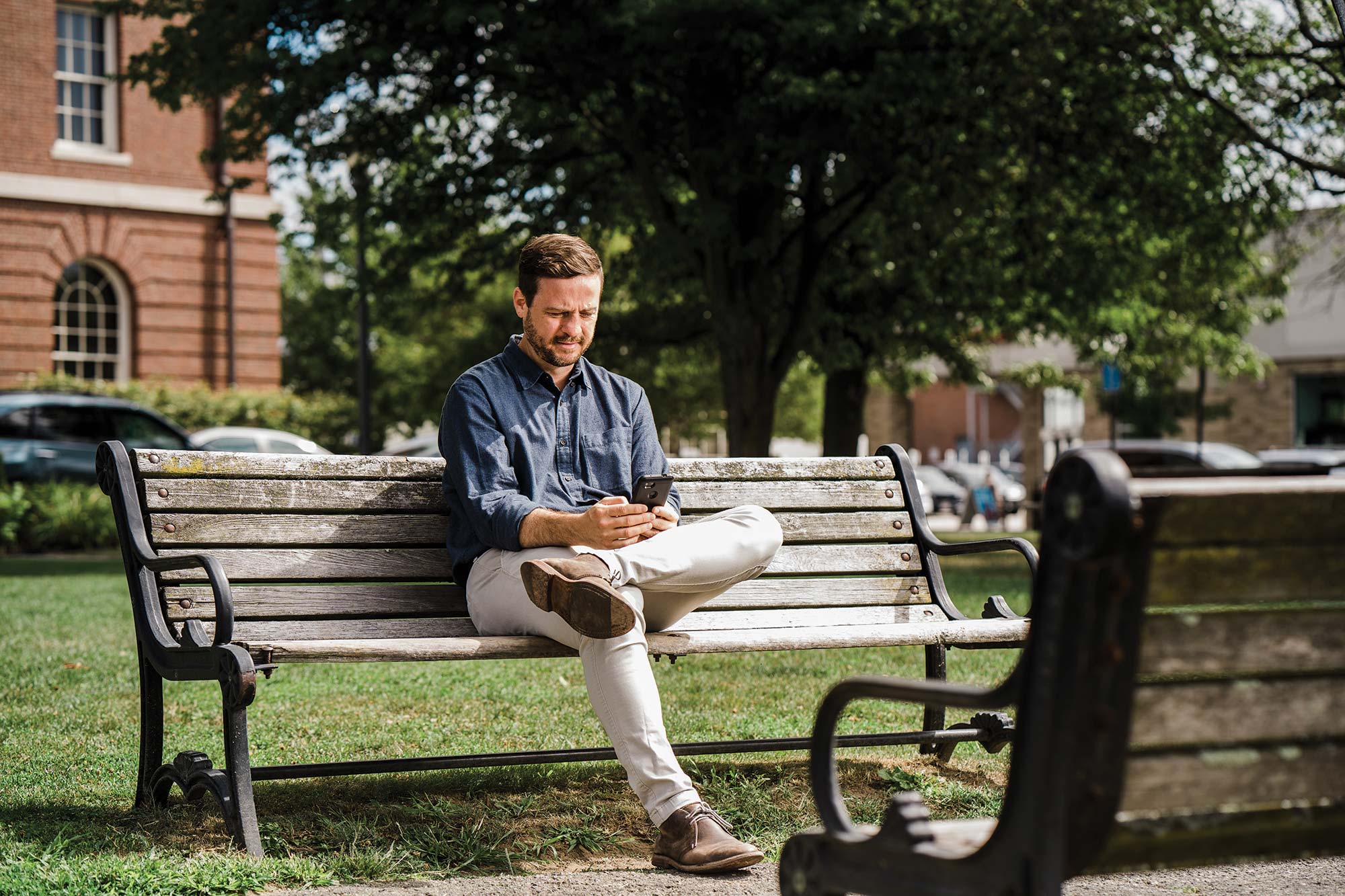 Photo of a young man sitting on a park bench by himself and looking at his phone.