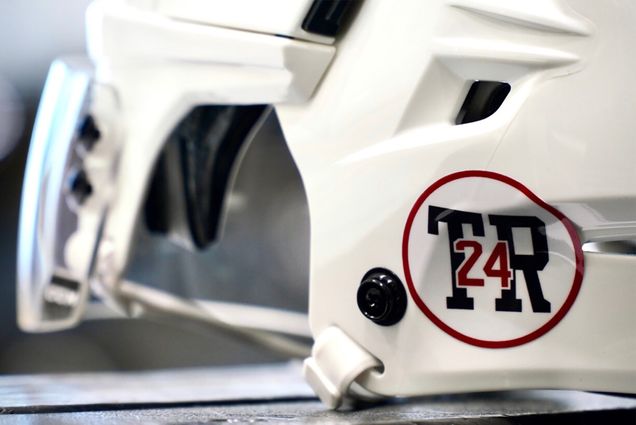 Closeup photo of a white hockey helmet with a special logo in honor of Travis Roy. The logo has a black “TR,” foregrounded by the number “24" in red, surrounded by a red circle.