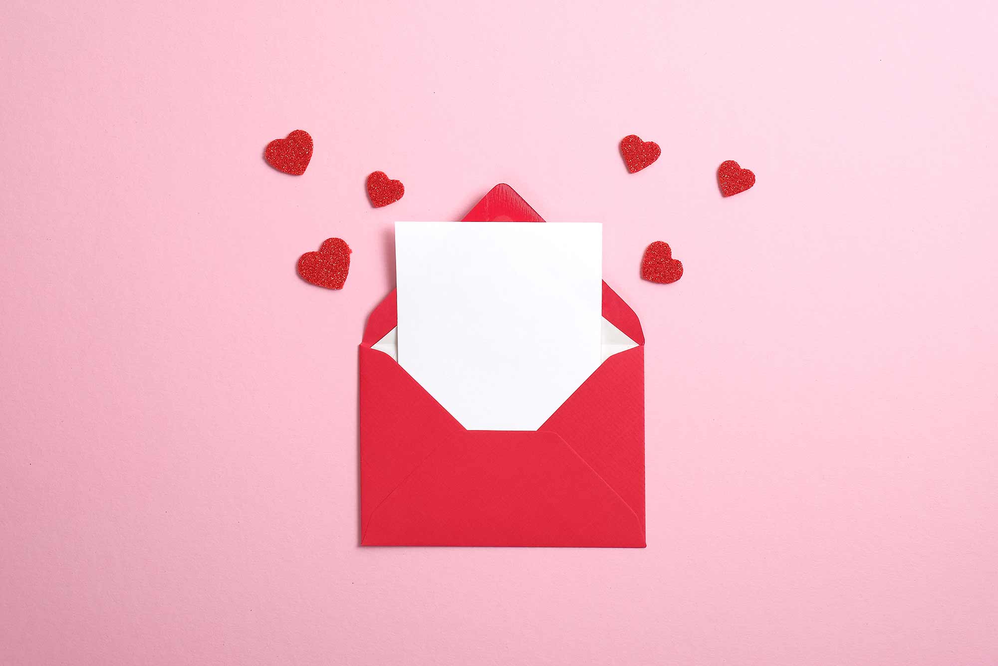 Blank Valentine's Day card in red paper envelope with blank white note mockup inside and Valentines hearts on pink background.