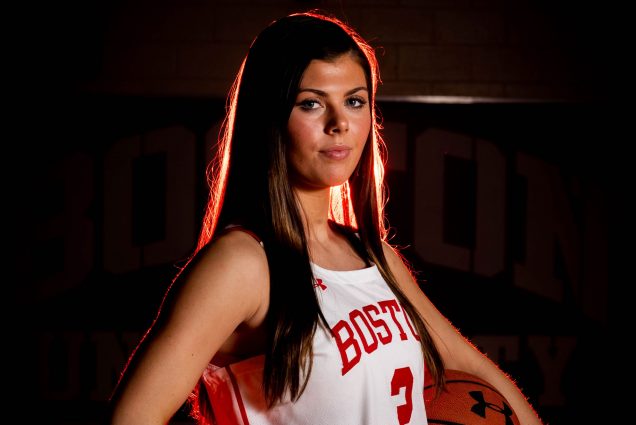 A portrait photo of Katie Nelson in her BU basketball uniform. She is standing with one hand on her hip, and the other holding a basketball.