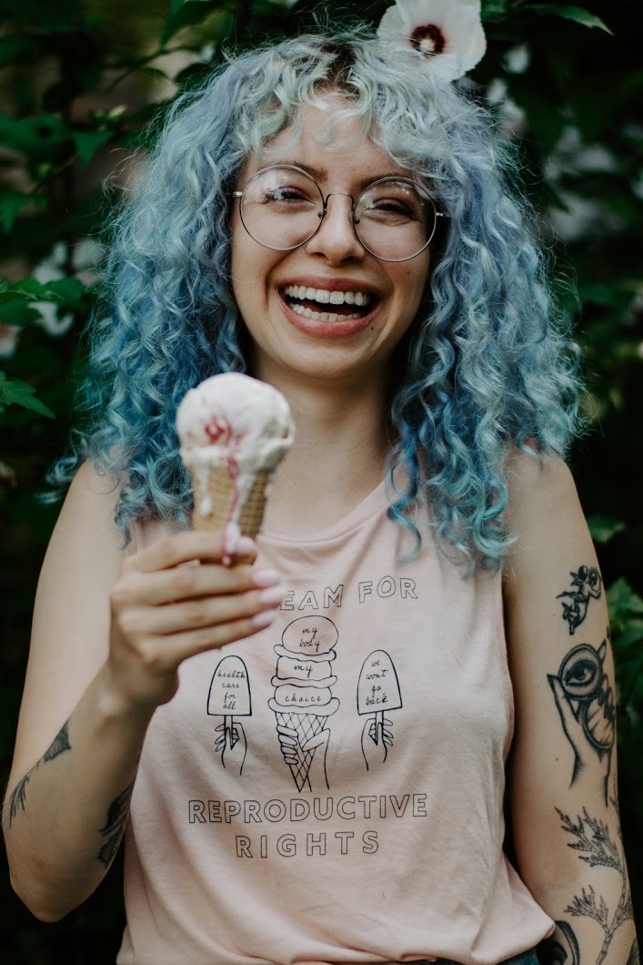 Photo of Hannah Spiegelman, a young woman with round glasses, icicle-blue curly hair, wearing a pink shirt with ice cream cones and the phrase “I scream for reproductive rights.” Hannah holds a dripping ice cream cone and smiles wide, as if mid laugh. A flower bush is seen blurred behind her.