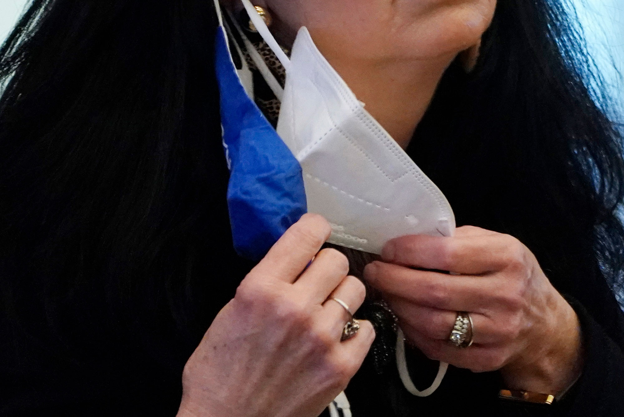 A photo of a woman with two masks on hanging off of one ear. She is holding the other end of both masks in her other hand.