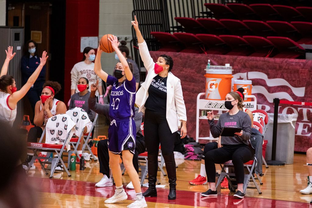 A photo of coach Maria Moseley on the sideline during a game. She is wearing a mask, standing with her hand up.