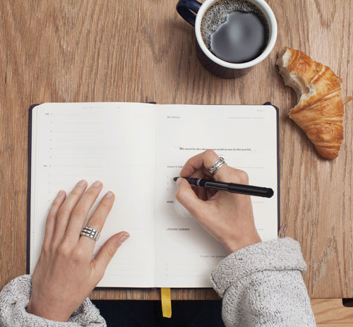 A photo of a persons hands writing in a notebook. Above the notebook are a cup of black coffee and a croissant with a bite taken out of it.