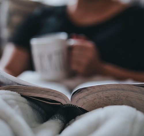 A photo of someone sitting under a blanket holding a cup of coffee while reading a book