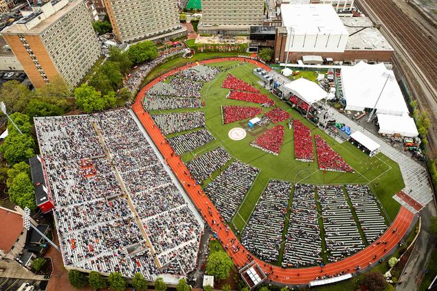 Aerial view of the 2019 Boston University Commencement on Nickerson Field on May 19, 2019. On the field, there are triangle and rectangular groups of people dressed in red on the right side of the field, then groups of white chairs, and gray bleachers furthest left.