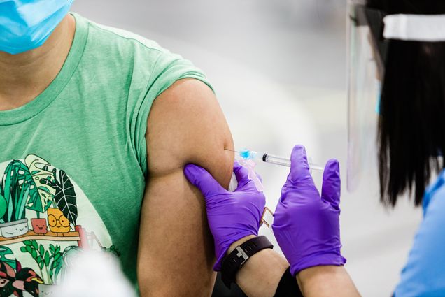 Photo of a medical professional in a face shield, blue mask, purple gloves, and a blue shirt giving a person in a green t-shirt a COVID vaccine in their arm. The photo is zoomed in and neither person’s face is seen.