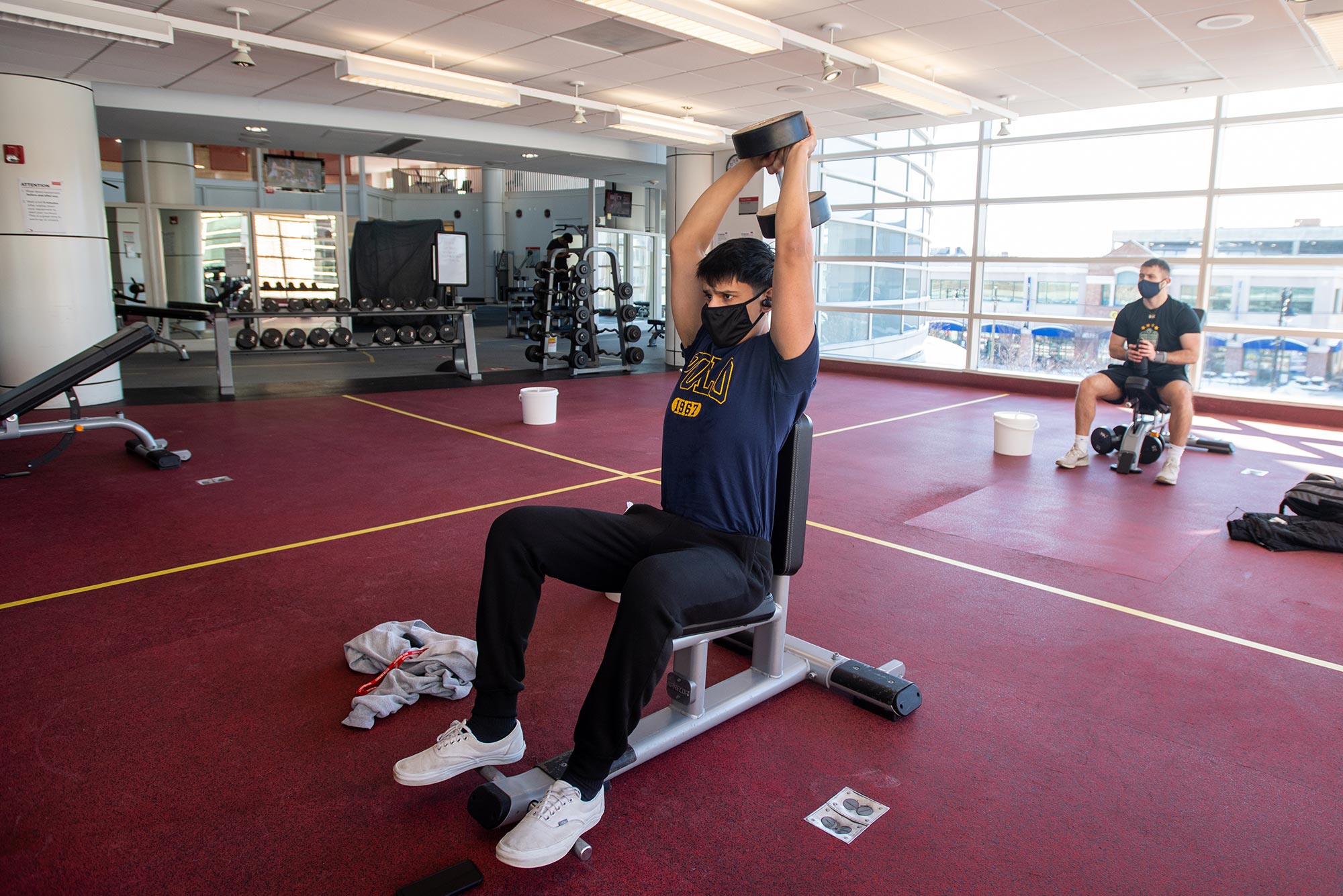 Photo of Shazan Khan (CAS’24) in a blue t-shirt and black sweatpants as he does an exercise in which he raises a barbell behind his head; another person working out a FitRec is seen seated behind him and a wrack of barbells are seen along the mirrored far wall.