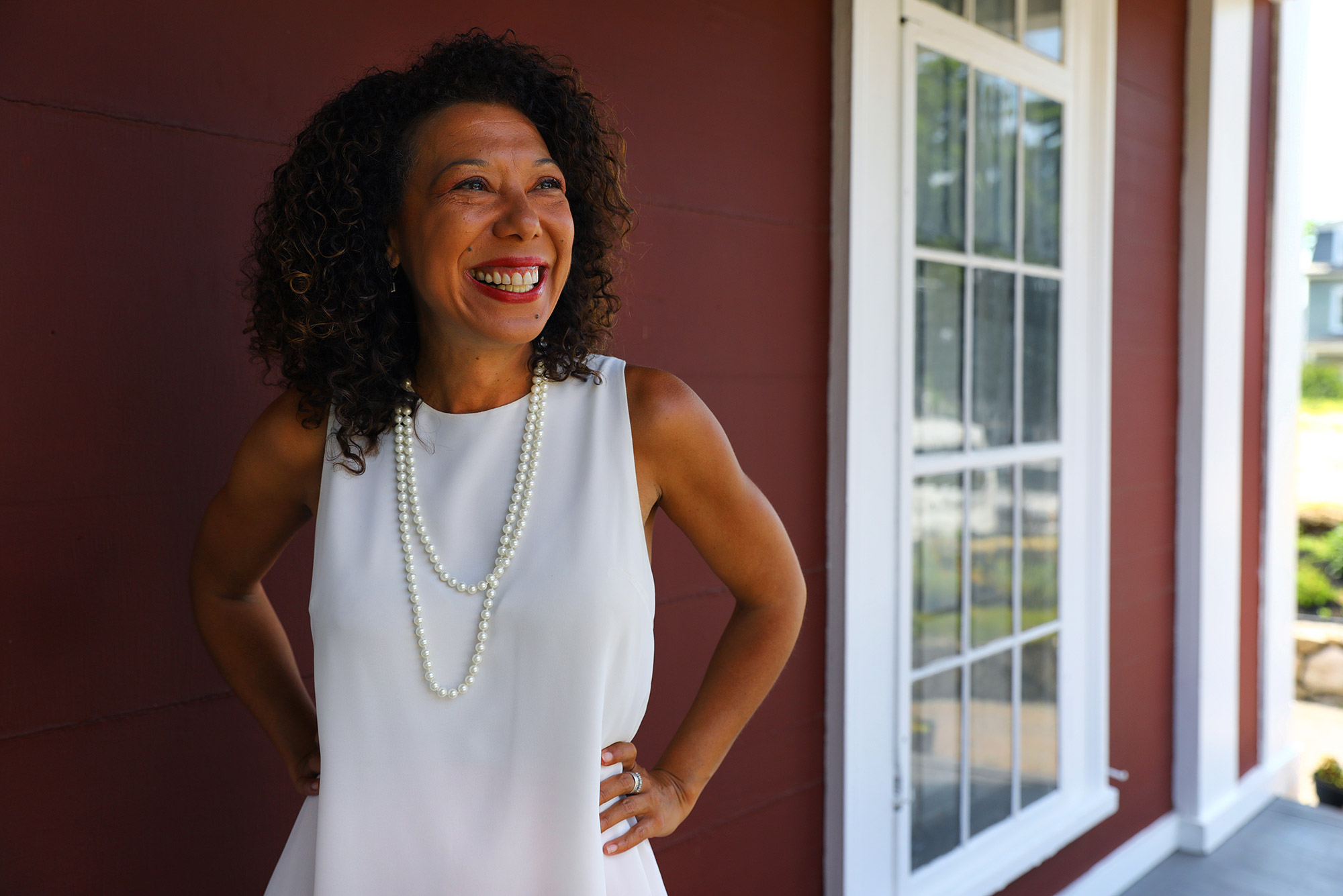 Photo of diversity advocate and organizer Malia Lazu in a white sleeveless blouse and pearls looking off to her left with her hands on her hips as she smiles wide. Behind her, a maroon wall and white window is seen.