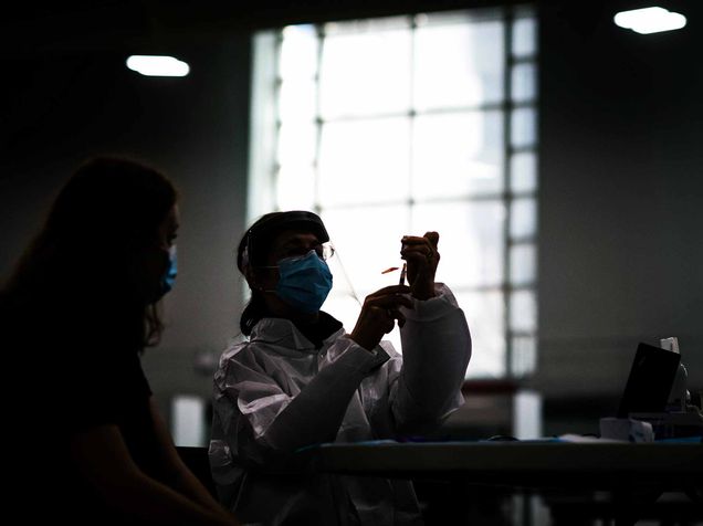 Photo of director of Nursing, Anne Patey, preparing to give a patient the Covid vaccine on January 15, 2020. The shot is very dark, and Patey is seen in silhouette holding the vaccine at eye-level while seated. She wears a face mask and face shield, and sun light comes in from a window behind her.