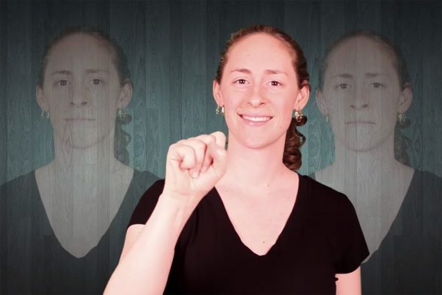 Photo of Elsie Stecker signing in ASL during a video on the word 'commotion.' Elsie wears a black v-neck and smiles as she seems to make the sign for the letter 'M'
