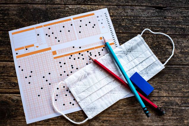 Photo of an orange and white multiple choice sheet resting on a desk, with some pencils and a blue face mask on top.