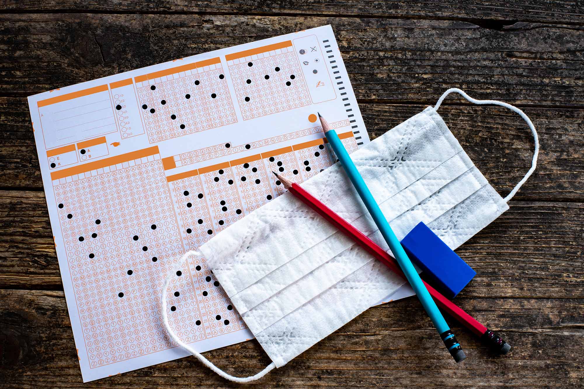 Photo of an orange and white multiple choice sheet resting on a desk, with some pencils and a blue face mask on top.