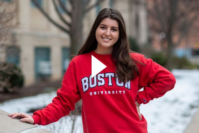 Photo of Vlogger Anna Malygin (Questrom'23), smiling, with her hand resting on a waist-high wall. She wears a bright red Boston University Sweatshirt, and has her hair down. Behind her, a blurred wall of CAS is seen, snow is on the ground. White play button is overlaid.