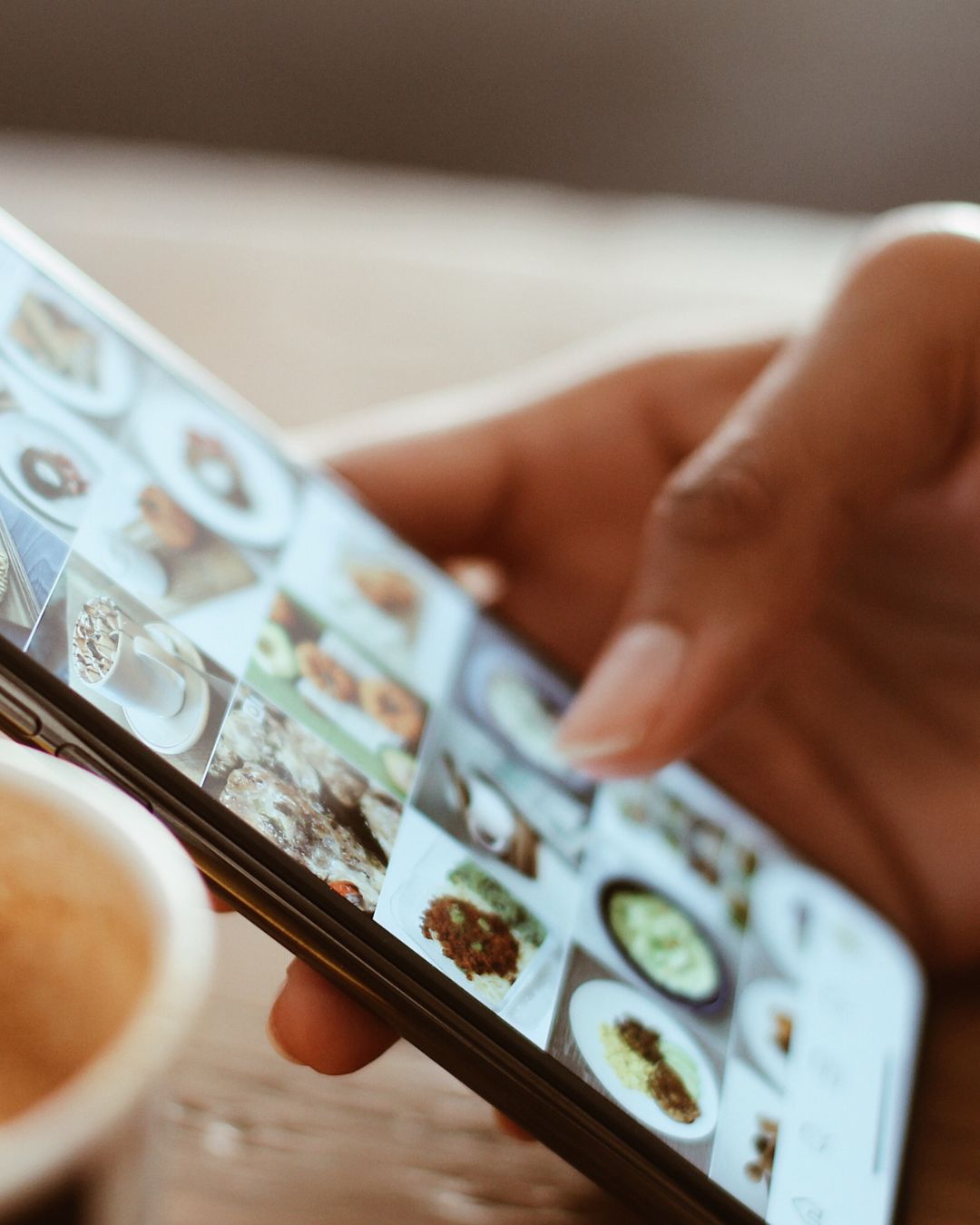 A hand with tan skin scrolls a phone screen full of pictures of food next to a coffee mug.