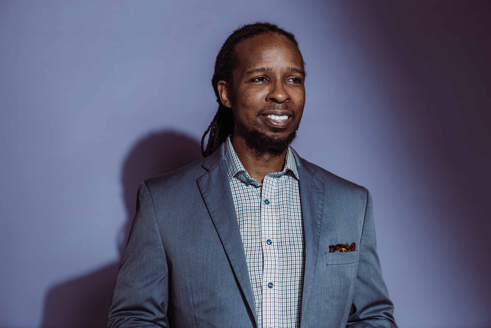 Portrait of Ibram X. Kendi standing in front of a lavendar background