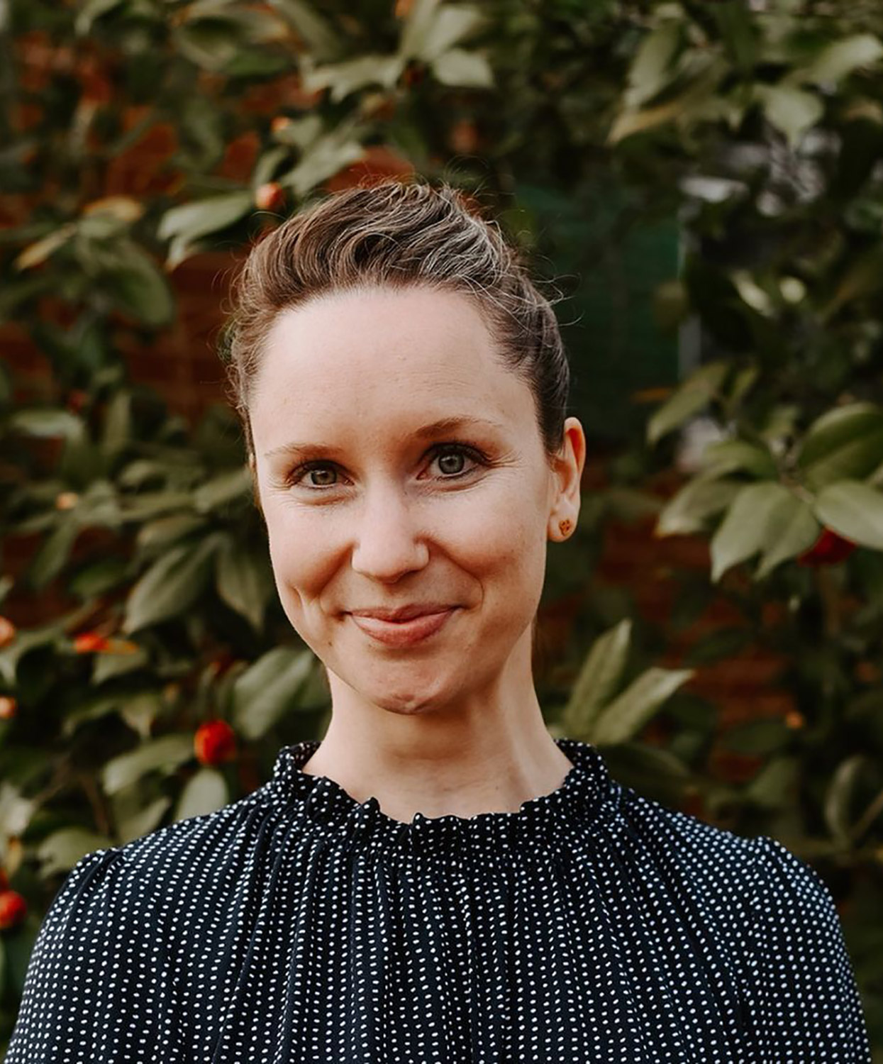 A portrait of Emily Contois, a young woman with her hair pulled back and a blue ruffled blouse smiles welcomingly. Behind her, a large push with red flowers is seen.