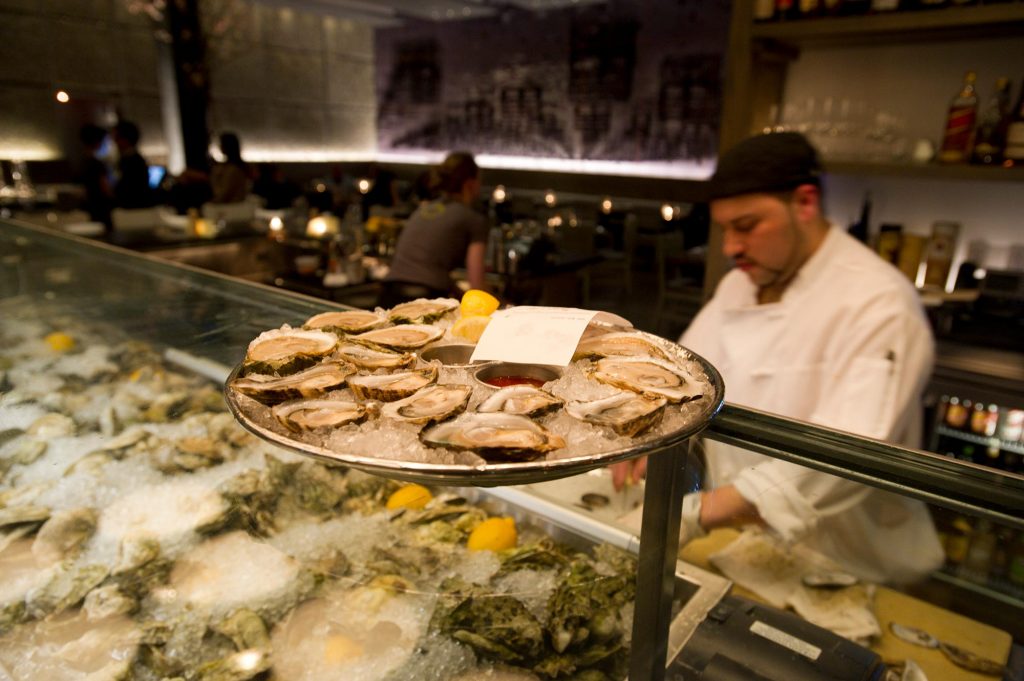 Photo of a tray of opened oysters with lemons and a ticket resting on the glass guard above a large ice cooler filled with unopened oysters. A line cook in a white jacket stands behind the counter opening oysters.