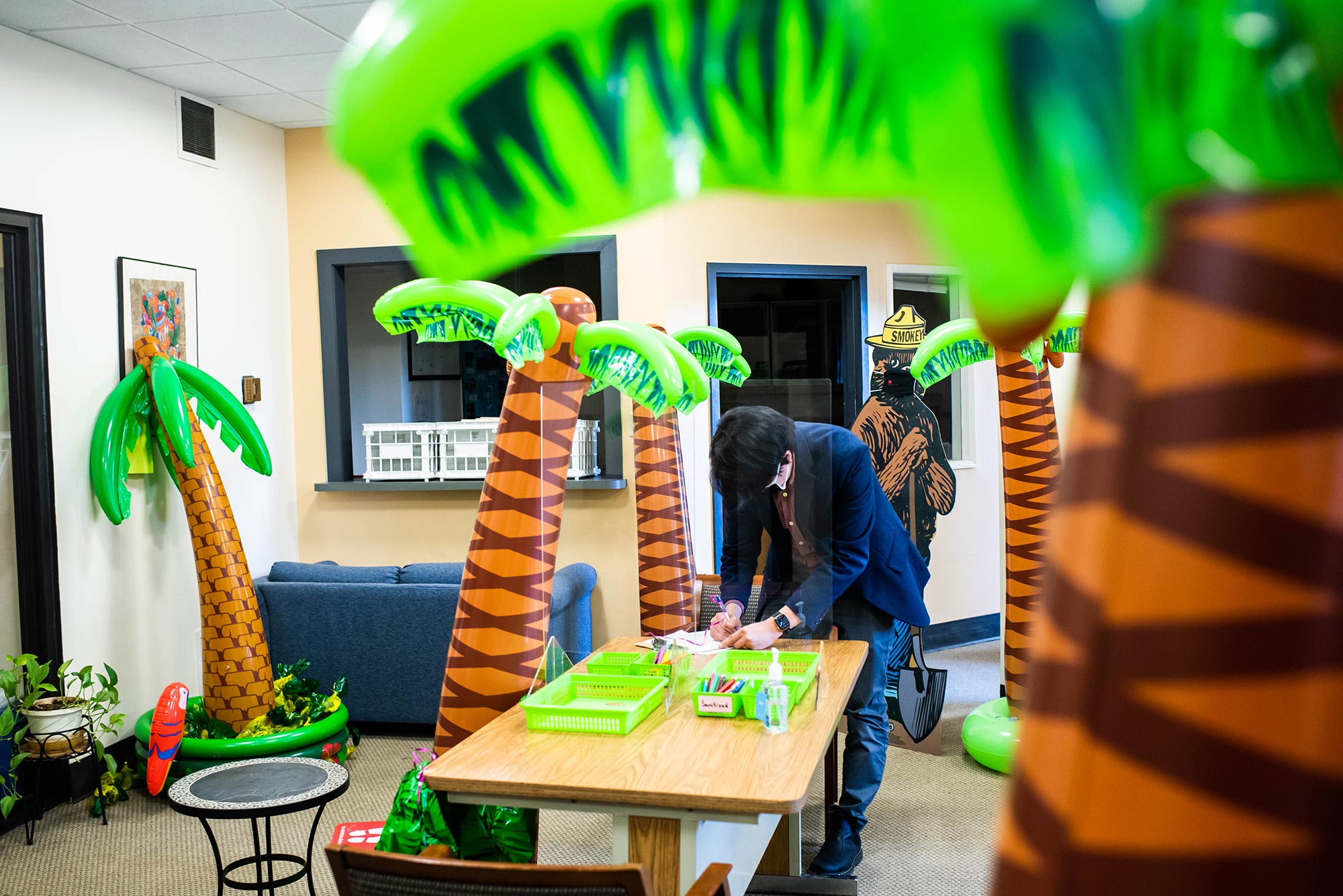 Photo of Sangwon Yang writing on a table at the center of the School of Theology’s registrar’s office, which is filled with 5 large, inflatable palm trees. Behind him, a Smokey the Bear cardboard cut out with a face mask on is seen.