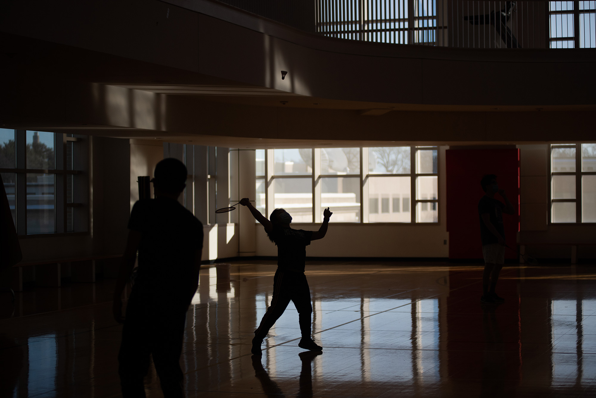 Photo of Chui Ting Poon (SAR’24), in silhouette, reaching back with a badminton racket to hit a shuttle cock, at Fitrec as she plays badminton with her club team. Light pours in through the windows towards the back of the gym and shines on the floor. Someone in silhouette is also seen in the front foreground.