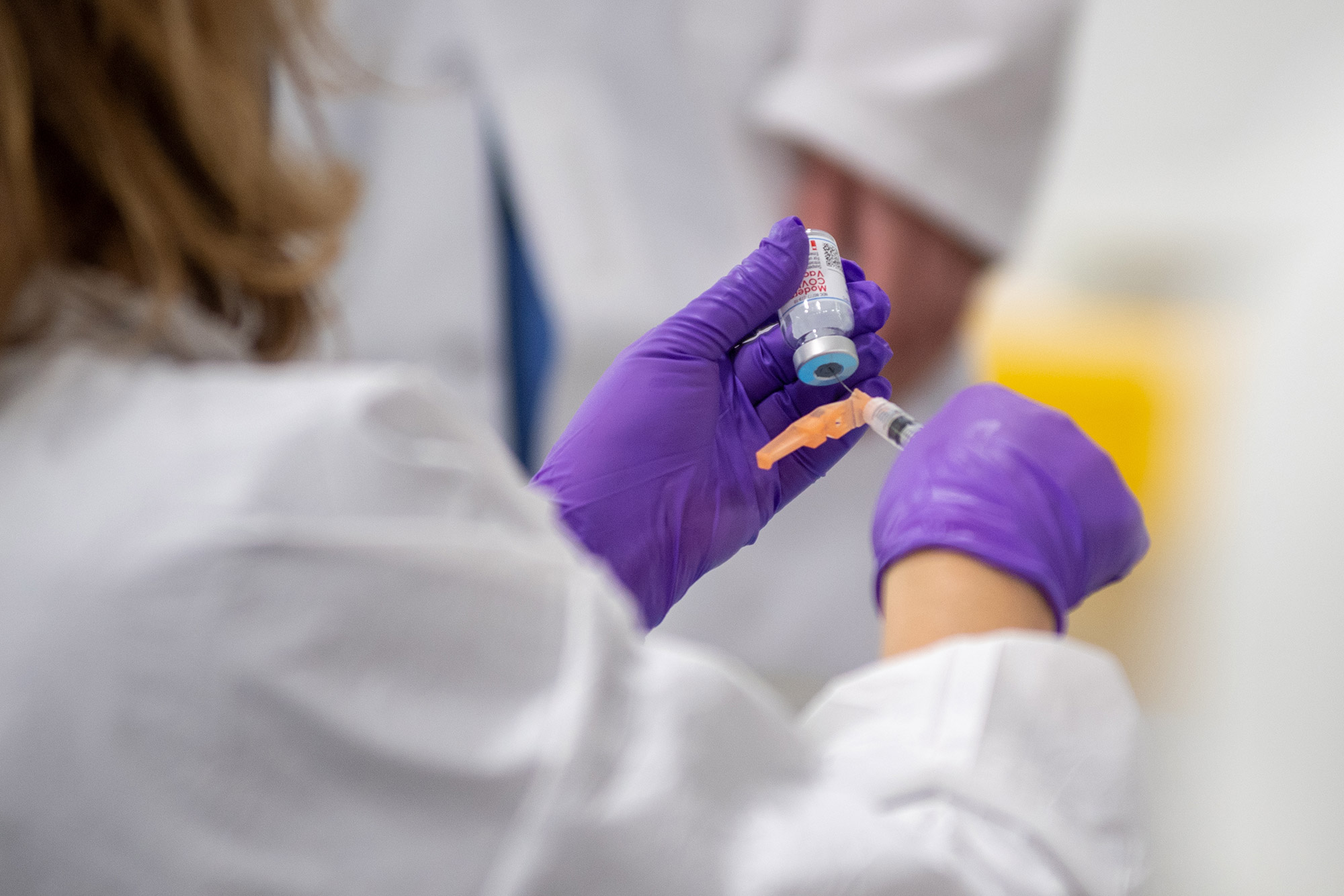 Photo of a pair of hands with purple gloves on extracting vaccine from a vial with a syringe.