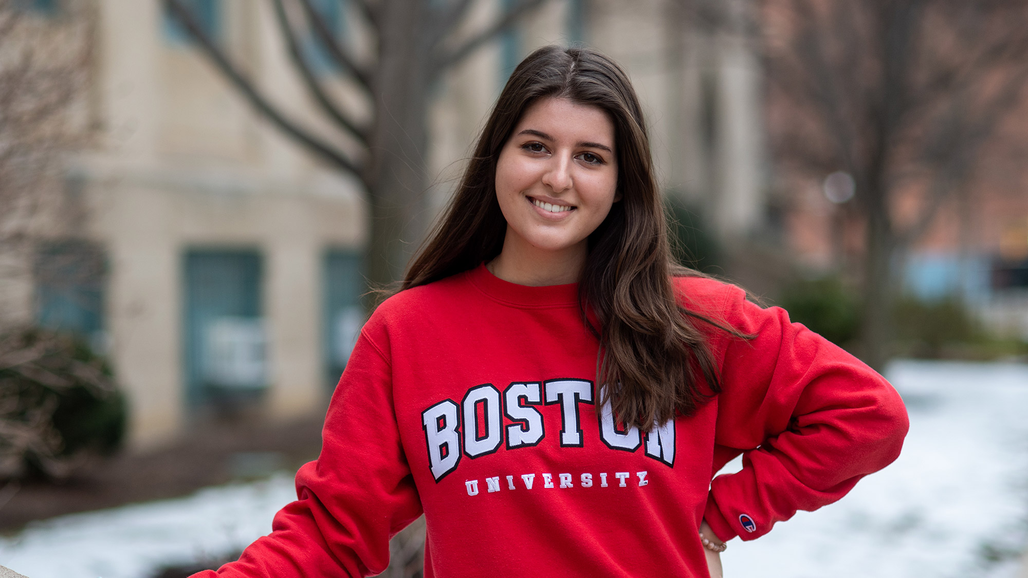 Photo of Vlogger Anna Malygin (Questrom'23), smiling, with her hand resting on a waist-high wall. She wears a bright red Boston University Sweatshirt, and has her hair down. Behind her, a blurred wall of CAS is seen, snow is on the ground.