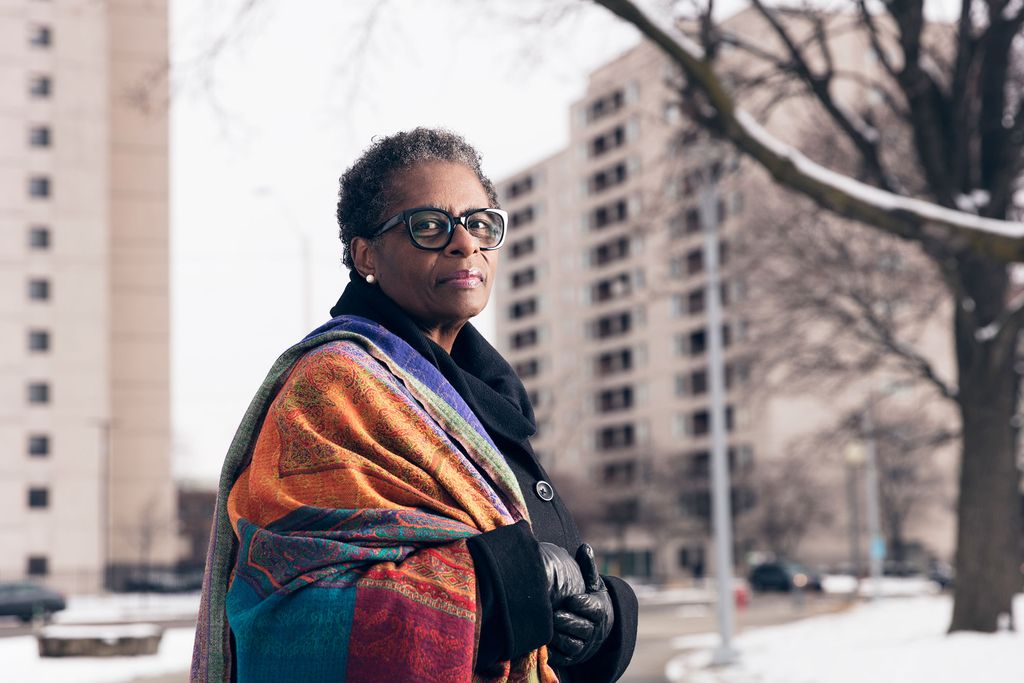 Photo of Henriquez, a middle-aged Black woman, standing in a long p-coat and colorful scarf in a snowy park. A large apartment building is seen behind her.