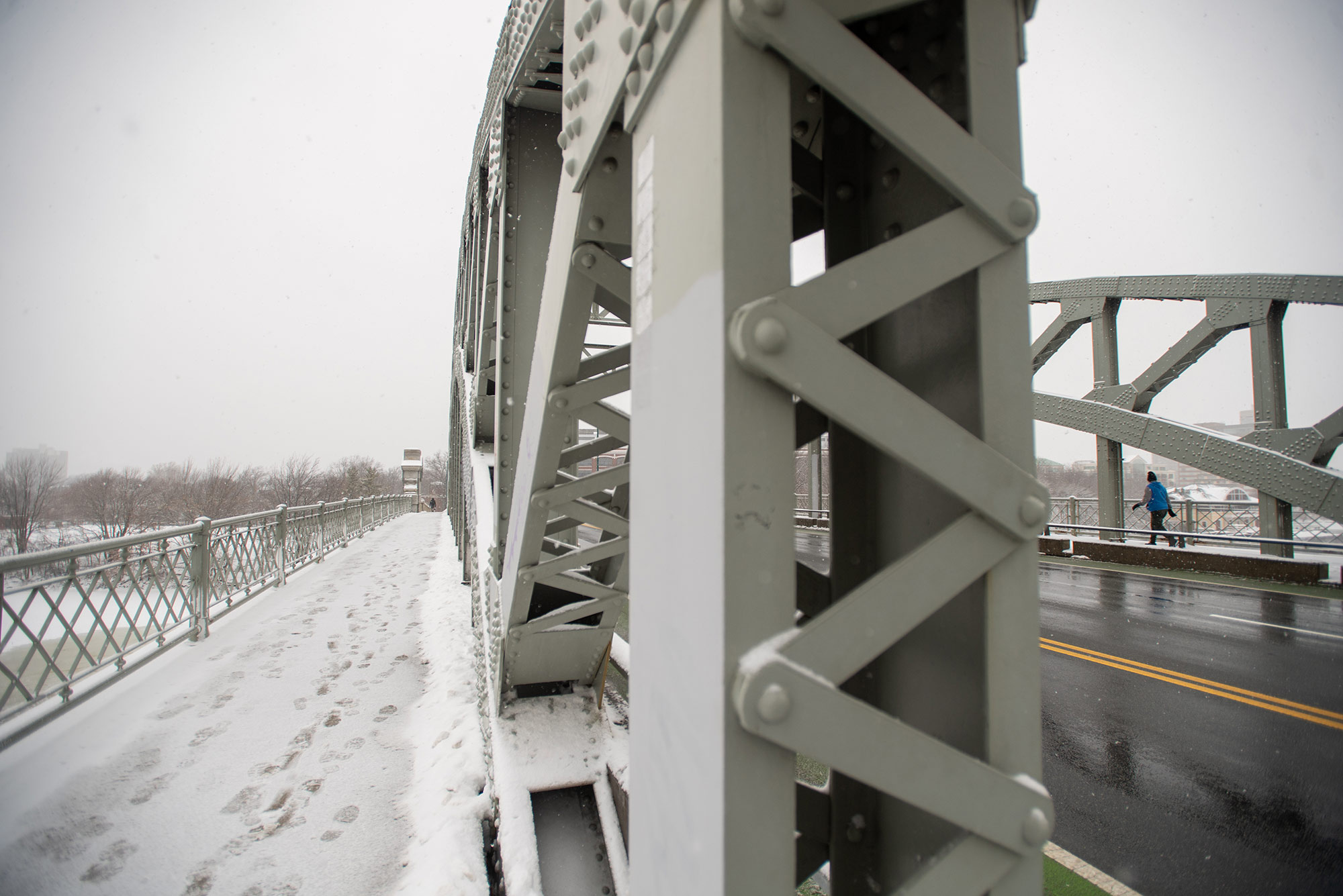 Photo taken on the BU Bridge, looking down the length of the bridge towards the Cambridge side. Footprints in the snow are seen on the walkway; the photo is foreground by some of the metal cross beams of the bridge.