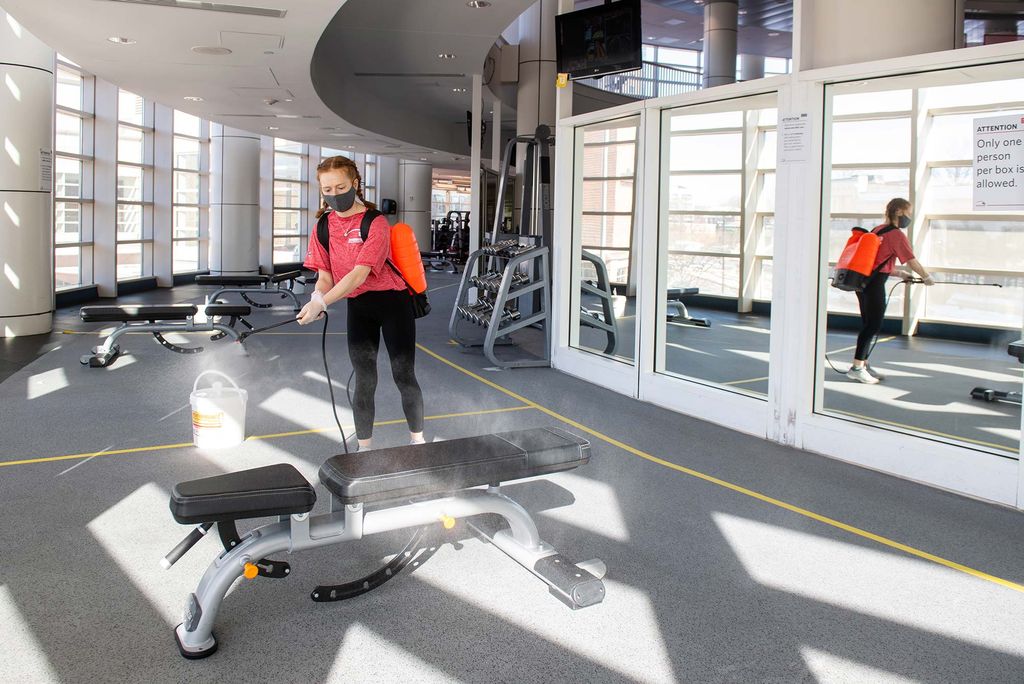 Lauren Gerlin (WHEELOCK’21), who works at FitRec, in a light red shirt, as she sprays down the workout stations between reservations at Fitrec. She wears a backpack of the fluid on her back and holds a sprayer. 